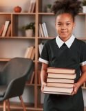 Holding books in hands. Cute african american girl in school uniform is at home library
